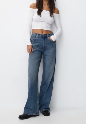 Femme portant un crop top blanc à manches longues et épaules dénudées, un jean bleu taille haute à jambes larges et des chaussures noires, debout devant un fond blanc.