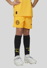 Uniforme de fútbol juvenil que incluye una camiseta amarilla y pantalones cortos, calcetas negras con letras amarillas, y botas blancas con detalles grises.