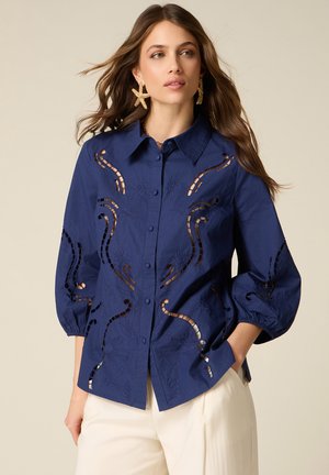 Kvinde med langt hår iført marineblå knapbluse med udskåret broderi og søstjerne-øreringe, kombineret med hvide bukser.