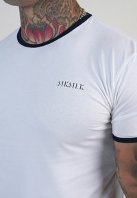 Balta trumpa rankovė apvalios iškirptės marškinėliai, savaime laikanti mėlynos spalvos kakliuko apdaila ir subtilus "SIKSILK" logotipas krūtinėje. Lygi medžiagos tekstūra.