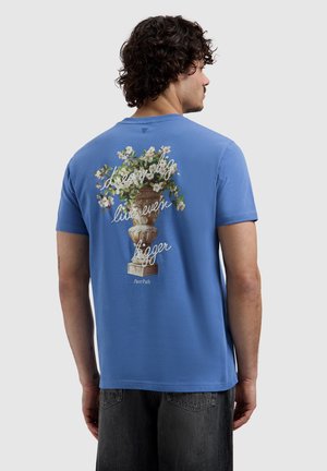 Man met krullend haar draagt een blauw T-shirt met een met bloemen gevulde stenen urn en de tekst 'droom groot, leef nog groter' op de achterkant.