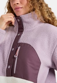 Polarowy pulower w jasnym lawendowym kolorze z ciemnofioletowym panelem z przodu, z zamykaną kieszenią i zatrzaskami na kołnierzu.