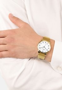 Gouden metalen polshorloge met een witte ronde wijzerplaat, zwarte cijfers en een gestructureerde mesh band, gedragen om een hand die op een wit shirt rust.