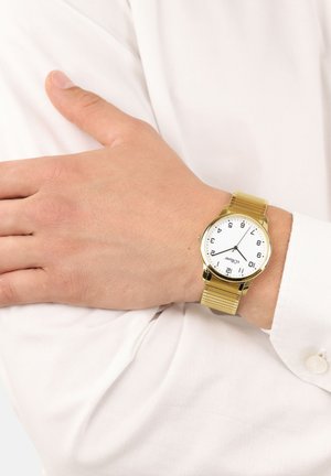 Goldfarbene Metallarmbanduhr mit einem weißen, runden Ziffernblatt, schwarzen Ziffern und einem strukturierten Mesh-Armband, getragen an einer Hand, die auf einem weißen Hemd ruht.
