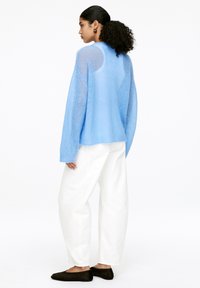 Frau mit lockigem Haar, die einen hellblauen Strickpullover, weiße Hosen mit weitem Bein und schwarze Slipper trägt, seitlich auf einem weißen Hintergrund stehend.