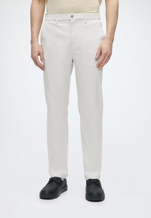 Homme portant un pantalon beige slim-fit avec fermeture par bouton et fermeture éclair, assorti à des chaussures noires à lacets sur un fond blanc uni.