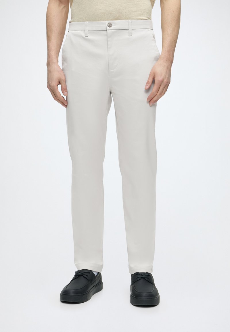 Homme portant un pantalon beige slim-fit avec fermeture par bouton et fermeture éclair, assorti à des chaussures noires à lacets sur un fond blanc uni.
