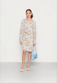 Vestido floral azul claro com mangas longas, cintura cingida e bainha até ao joelho. Combinado com uma bolsa texturizada azul claro e sandálias de cor creme.