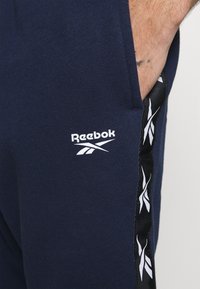 Tmavě modré jogger kalhoty vyrobené z měkké látky, s bílým logem Reebok a černým bočním pruhem s bílým akcentním designem.