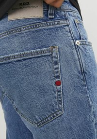 Plave traperice s istaknutim stražnjim džepom, kontrastnim crvenim šivom i kožnom etiketom označenom "R.D.D. Royal Denim Division."