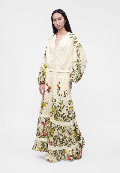 Femme aux longs cheveux raides portant une longue robe crème à motif floral, manches volumineuses et détails en dentelle, debout sur fond blanc.