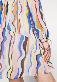 Robe longue à manches longues en tissu transparent avec des rayures ondulées pastel en bleu, rose, jaune, marron et noir, nouée au poignet avec un nœud.