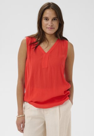 Kobieta o długich brązowych włosach, ubrana w bezrękawnik czerwony top z dekoltem w serek oraz jasnobeżowe spodnie, stoi z jedną ręką w kieszeni na jednolitym tle.