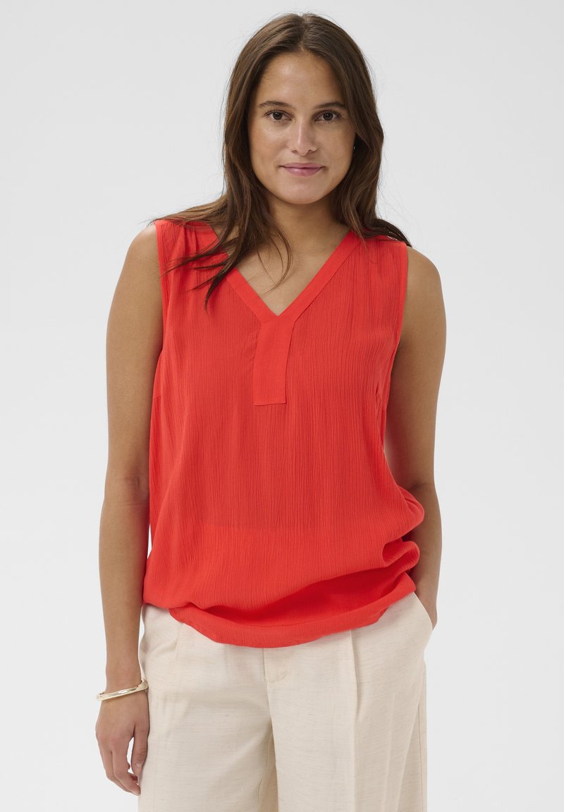 Femme aux longs cheveux bruns portant un haut rouge sans manches à col en V et un pantalon beige clair, debout avec une main dans la poche devant un fond uni.