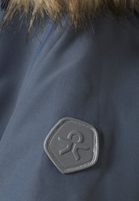 Mörkblått tyg har en texturerad yta med en grå hexagonal patch som visar en symbol, i kombination med brun pälskant längs kanten.