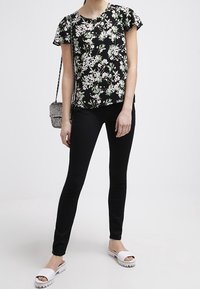 Černý květinový top s krátkými rukávy, v kombinaci s černými úzkými kalhotami a bílými pantoflemi s texturovanou podrážkou. Malá kovová kabelka.