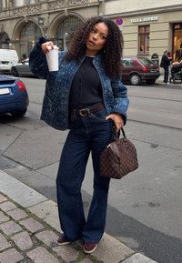 Kurtka dżinsowa z wzorem paisley, czarny top, wysokie dzwony z podwyższonym stanem, brązowa torebka w kratę, trzymająca kubek kawy, swobodna miejska sceneria.