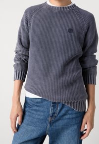 Person, der einen strukturierten grauen Strickpullover mit gerippten Bündchen und Saum über blauen Jeans trägt, vor einem schlichten hellen Hintergrund steht.