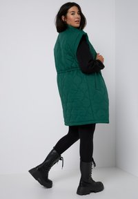 Grön vadderad väst med bälte i midjan, hög krage och ärmlös design, ihop med svarta leggings och kraftiga svarta stövlar.