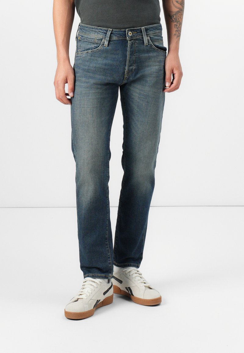 Jeans de mezclilla azul oscuro con corte recto, textura desgastada, cinco bolsillos y cierre de botón, combinados con zapatillas blancas.