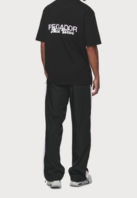 Černé bavlněné tričko s potiskem "PEGADOR Next Wave" na zadní straně; spárované s černými kalhotami s růžovými bočními pruhy a elastickými manžetami.