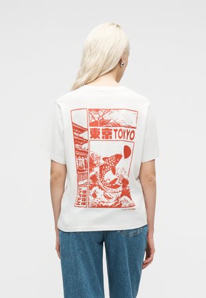 Kvinde med langt blondt hår iført hvid T-shirt med rød grafik af koi-fisk, Mount Fuji og japansk tekst, parret med blå jeans.