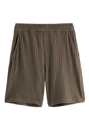 Dreng iført tekstureret brun kortærmet skjorte med knapper og matchende shorts, hvide sokker og sorte sneakers, stående mod en ensfarvet baggrund.
