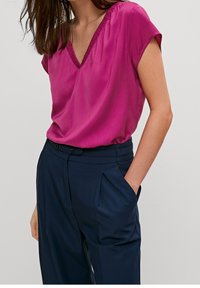 Blouse à manches courtes fuchsia avec un col en V et un col froncé, associée à un pantalon taille haute bleu marine avec des plis et des poches latérales.