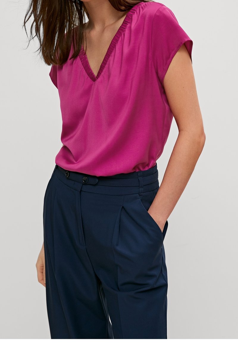 Blouse à manches courtes fuchsia avec un col en V et un col froncé, associée à un pantalon taille haute bleu marine avec des plis et des poches latérales.