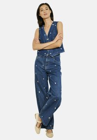 Sieviete stāv ar rokām sakrustotām, ģērbusies zilā džinsu veste un biksēs ar maziem baltiem ziedu izšuvumiem, valkā beigtas plakanās kurpes.