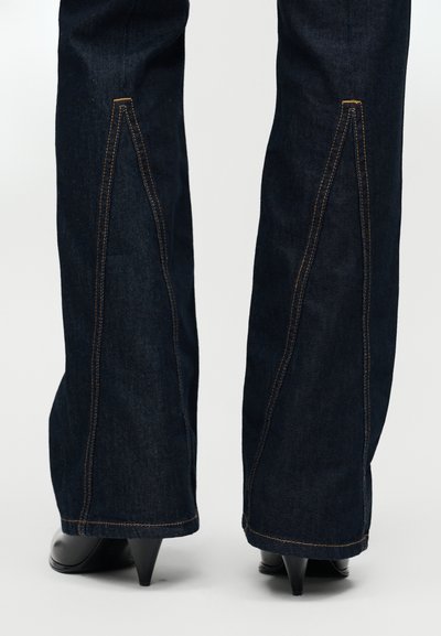 Σκούρο τζιν καμπάνα με έντονες αντίθετες ραφές σε πορτοκαλί χρώμα, με διακριτική λεπτομέρεια ραφής κατά μήκος των εξωτερικών ποδιών. Εφαρμοστό γύρω από την κορυφή, που φαρδαίνει προς τα κάτω.