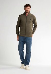 Jachetă tricotată verde măsliniu cu fermoar, cu guler înalt, prevăzută cu o mică aplicație neagră și manșete striate; asortată cu blugi albaștri și adidași albi.