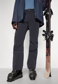 Skihosen aus dunkelgrauem Stoff, mit geradem Schnitt und verstärkten Knien. Schwarze Skistiefel mit verstellbaren Schnallen und ein Ski mit schwarzen Bindungen.