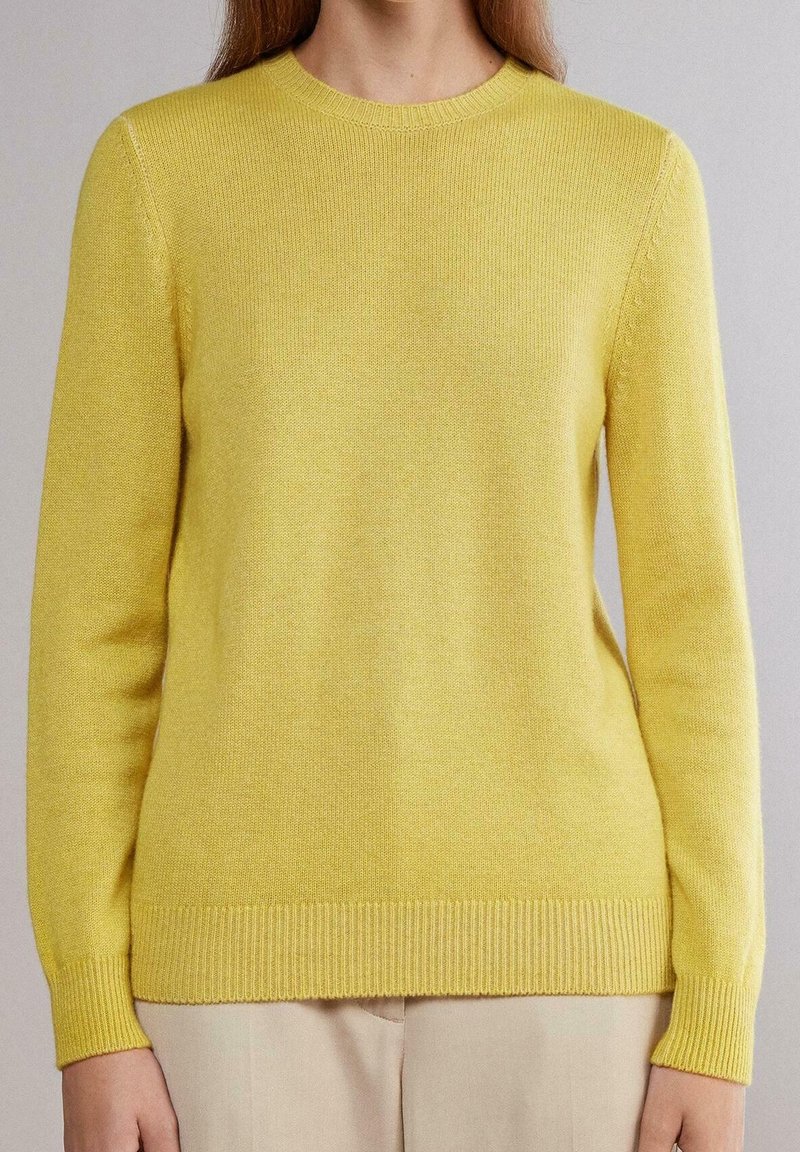 Femme portant un pull en tricot jaune uni à manches longues avec des poignets et un ourlet côtelés, associé à un pantalon beige clair sur un fond neutre.
