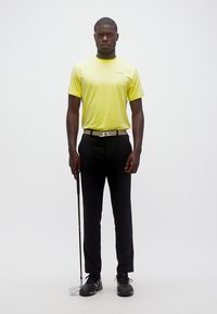 Άνδρας με κίτρινο πουκάμισο και μαύρο παντελόνι που κρατάει ένα μπαστούνι γκολφ, στέκεται μπροστά σε λευκό, απλό φόντο.