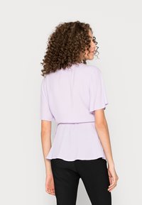 Blusa lilás de manga curta com cintura ajustada, textura suave e bainha fluida. Combinada com calças pretas, apresentando um corte descontraído.