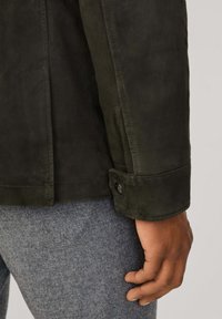 Giacca in suede verde scuro con dettagli di cucitura, dotata di colletto e polsini con bottoni, indossata sopra un tessuto grigio strutturato.