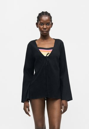 Femeie cu părul împletit, purtând un top negru cu mâneci lungi și decolteu în V peste un bandeau cu dungi multicolore, pe un fundal alb simplu.