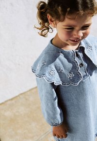Denimmekko vaaleansinisessä värissä, jossa on kukkakirjontaa ja hammastettu kaulus. Pitkät hihat, joissa on elastiset hihansuut.