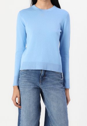 Femme portant un pull bleu clair à manches longues et un jean en denim bleu, debout devant un fond blanc.