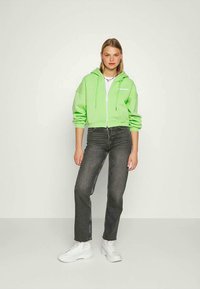 Hoodie verde claro cortado com cordões, fecho de zíper e texto impresso, combinado com jeans cinzentos de perna reta e ténis brancos.