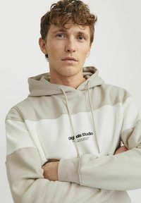 Beige og hvid hoodie med en stor kængurulomme, justerbar snøre ved hætten og "Originals Studio" logo på brystet. Glat stoftekstur.