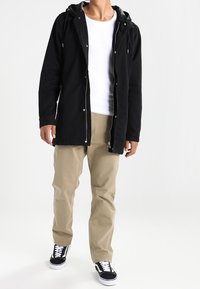 Giacca nera con cappuccio e zip frontale, abbinata a pantaloni beige e sneaker nere. Il tessuto sembra testurizzato con dettagli di cucitura visibili.
