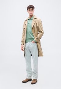 Jeune homme portant un trench-coat beige, un pull polo vert, un jean bleu clair et des bottes en daim marron, debout devant un fond blanc.