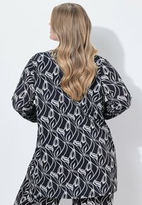 Volná černá tunika s bílým květinovým vzorem, dlouhými rukávy a pohodlným střihem. Tkanina vypadá hladce; design se táhne po celém oděvu.