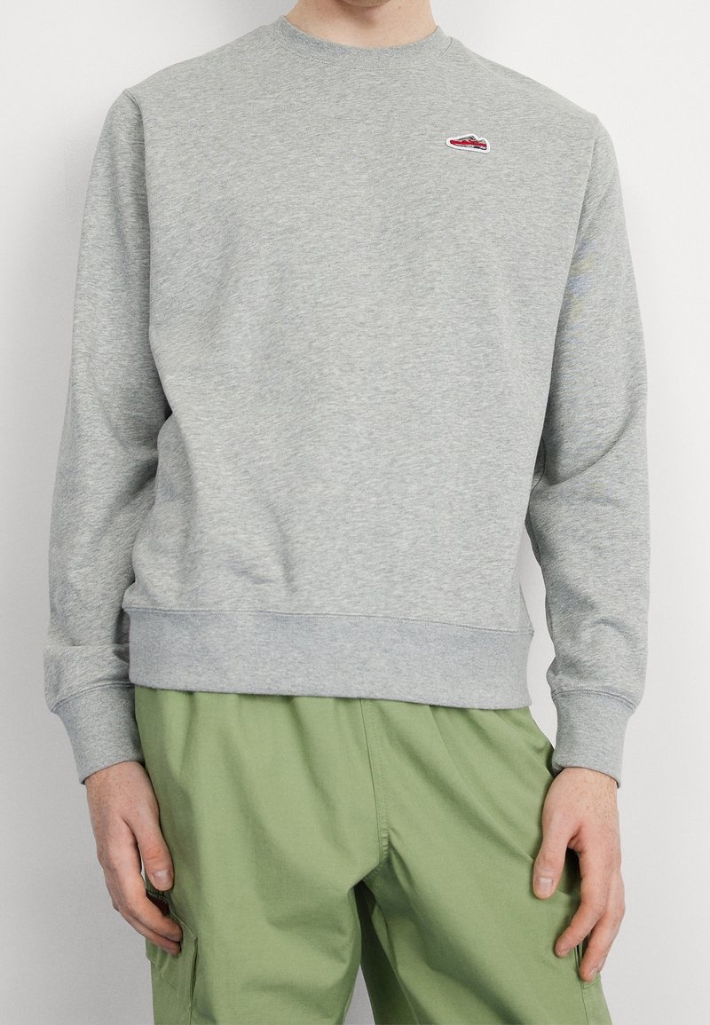 Grå sweatshirt med rund hals, ribbade muddar och fåll, med en liten röd broderad logotyp på bröstet. Matchas med gröna byxor.