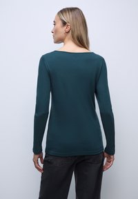 Teal langærmet top med en bred halsudskæring og glat tekstur, parret med mørke jeans. Stof er letvægts og let figursyet.