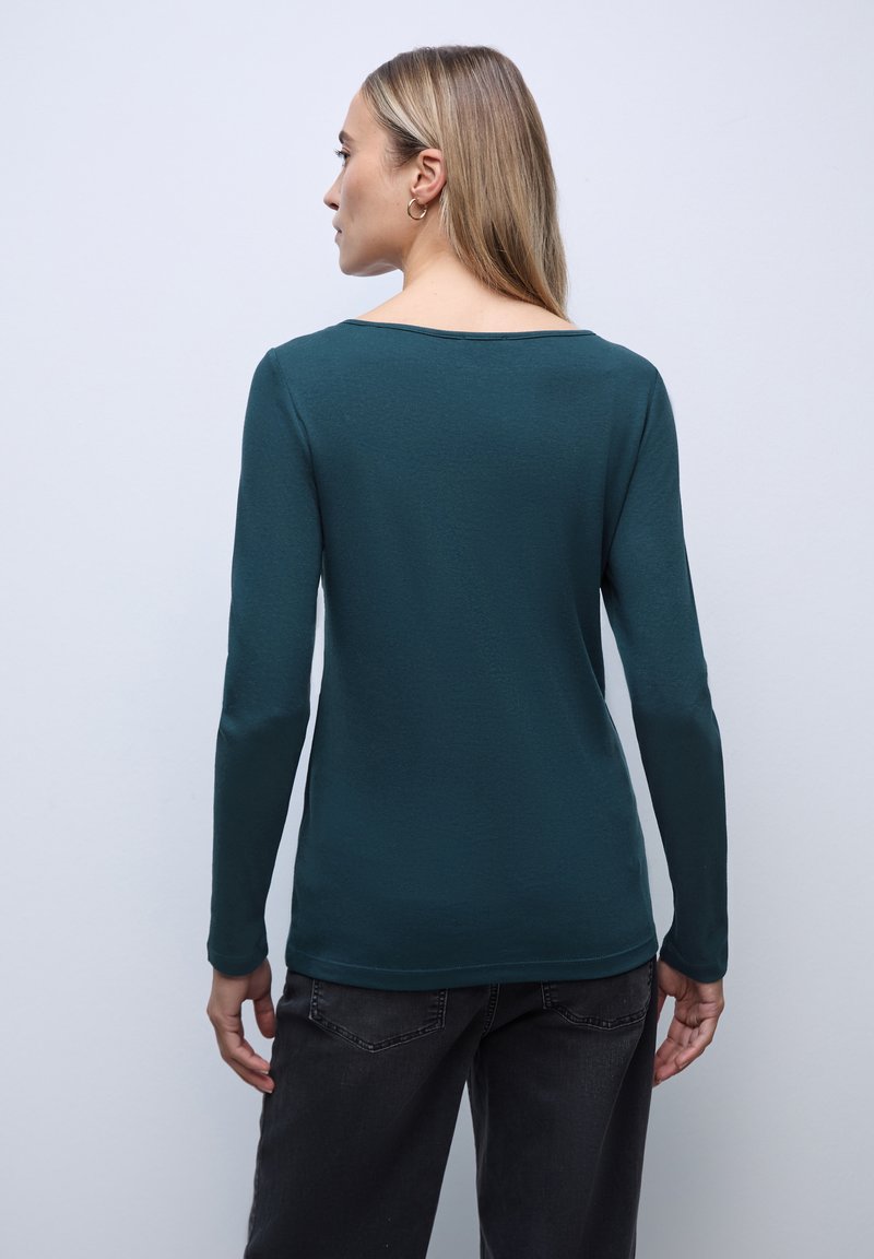Teal langærmet top med en bred halsudskæring og glat tekstur, parret med mørke jeans. Stof er letvægts og let figursyet.