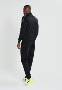 Ensemble de sport noir comprenant une veste à zip et un pantalon assorti. Fabriqué en tissu lisse avec des accents réfléchissants. Chaussures blanches et jaunes néon.
