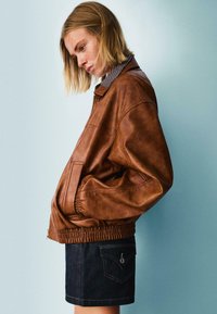 Giacca bomber in pelle marrone con vita elasticizzata, colletto stylizzato e tasche laterali, indossata con una mini gonna in denim scuro. Texture liscia.
